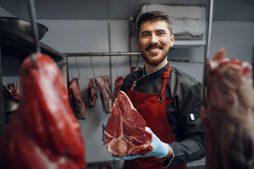 young butcher holding raw meat steaks in fridge of grocery shop